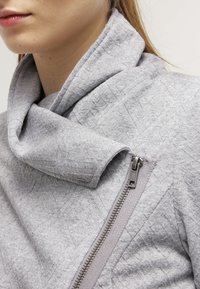 Šedý prošívaný top s nákrčníkem a asymetrickým zapínáním na zip. Vyrobený z texturované látky, s hladkým zipem a zakončenými okraji.