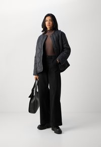 Črna prešita jakna s krožnim ovratnikom, rjav polo pulover, črne široke hlače in masivne črne platforme, v roki drži črno torbico.