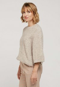 Pull en tricot léger beige, moelleux, avec un col rond et des manches courtes et larges. Porté avec un pantalon à carreaux marron et blanc.