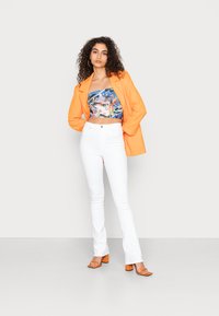 Blazer laranja sobre um top cropped de padrão colorido, combinado com calças brancas de boca de sino e sandálias de salto laranja. Fundo simples.
