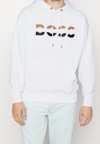 Sudadera blanca con un bolsillo canguro en el frente, capucha con cordón y el logo "BOSS" en negro, marrón y beige en el pecho.