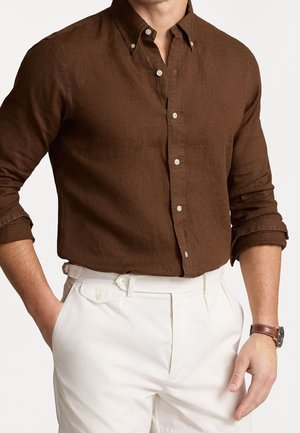 Homme portant une chemise marron à manches longues avec poignets retroussés et un pantalon blanc taille haute avec poche latérale et passants de ceinture.