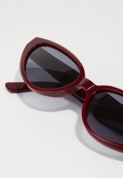 Burgundy sunglasses with large, oval frames and dark lenses. The frames are glossy with a smooth texture, featuring subtle branding on the inner arm.