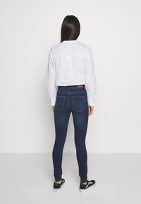 Jeans ajustados de denim oscuro, con una cintura de altura media, ajuste slim, costuras sutiles y dos bolsillos traseros. Combinados con una camisa blanca de botones.