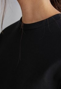 Schwarzes Sweatshirt mit Rundhalsausschnitt. Weiches Baumwollmaterial, glatte Textur und gerippter Kragen. Haarsträhnen sind am Kragen sichtbar.