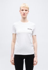 Άτομο με κοντά ξανθά μαλλιά, φορώντας άσπρο t-shirt με το κείμενο "AMOUR" και μαύρο παντελόνι, στέκεται μπροστά σε λευκό απλό φόντο.