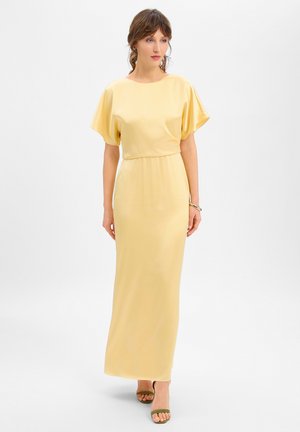 Frau trägt ein langes, hellgelbes Kleid mit kurzen Ärmeln, kombiniert mit olivgrünen offenen High-Heels, steht vor einem schlichten weißen Hintergrund.