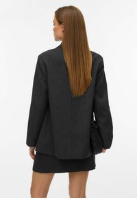 Žena s dlouhými rovnými rudými vlasy, oblečená v tmavém oversized blejzru a sukni, držící černou skládací kabelku, obrácená zády.