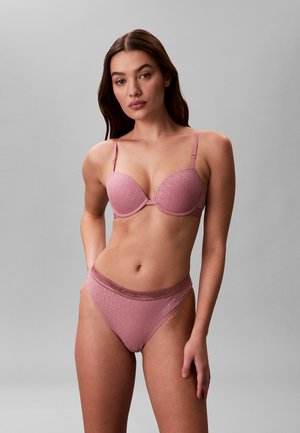 Frau mit langen braunen Haaren, die einen passenden pinken Calvin Klein BH und hochgeschnittene Unterwäsche trägt, steht vor einem grauen Hintergrund.