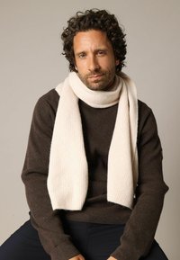 Écharpe beige côtelée portée par-dessus un pull en maille marron. L'écharpe a une texture douce et tombe librement autour du cou. Couleurs simples et neutres.