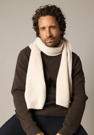 Écharpe beige côtelée portée par-dessus un pull en maille marron. L'écharpe a une texture douce et tombe librement autour du cou. Couleurs simples et neutres.