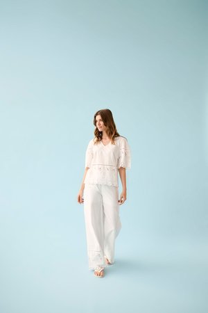 Nő sétál, fehér szemöldökös csipkeblúzát visel széles ujjakkal és hozzá illő bőszárú nadrággal, halványkék háttér előtt.