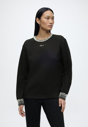 Donna con lunghi capelli neri che indossa un maglione nero con dettagli di perle sullo scollo e sui polsini, in piedi davanti a uno sfondo bianco semplice.