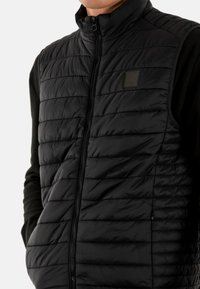 Zwart gewatteerd vest met hoge kraag, met horizontale doorgestikte textuur, ritssluiting aan de voorkant en subtiel logo op de borst. Lichtgewicht materiaal.