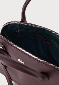 Bordeaux leren handtas met een ritssluiting, binnenste ritszak en Lacoste-logo. Gladde buitenkant met contrasterende donkere voering.