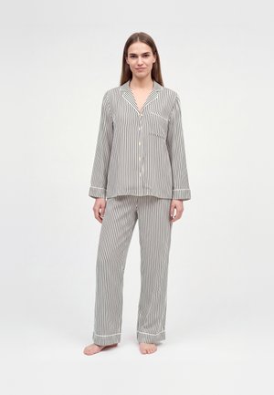 Une femme présente un ensemble de pyjama rayé assorti, composé d’une chemise boutonnée sur le devant et d’un pantalon ample. Elle se tient pieds nus devant un fond blanc uni.