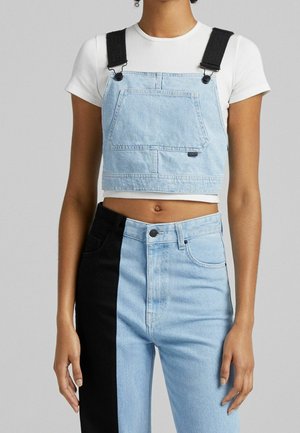 Persoon die een wit cropped T-shirt draagt, een lichtblauw denim cropped overall top en tweekleurige lichtblauwe en zwarte jeans.