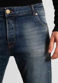 Tummansiniset farkut, joissa haalistuneita kohtia ja kulutettuja yksityiskohtia, oranssilla ompeleella, metallinapilla ja sivutaskuilla.