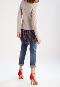 Beżowa dopasowana marynarka, czarna koszula w kropki nałożona warstwowo, niebieskie jeansy z podwiniętymi nogawkami, czerwone sandały na wysokim obcasie z paskiem wokół kostki, gładkie tekstury.