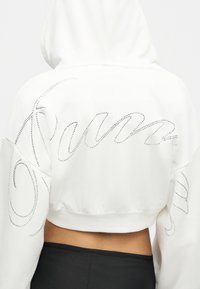 Οπίσθια όψη ενός ατόμου που φοράει λευκό cropped φούτερ με λεπτομέρειες από στρας σε καλλιγραφική γραφή και μαύρο παντελόνι.