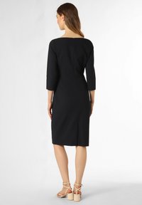 Schwarzes Kleid mit Dreiviertelärmel, quadratischem Ausschnitt und einem Reißverschluss im Rücken. Der Stoff wirkt glatt und hat eine figurbetonte Silhouette.
