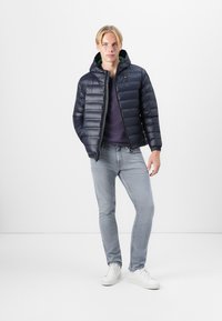 Navy puffer jacket with a zip closure and hood, featuring quilted design and elastic cuffs, worn over a gray sweater and light jeans.