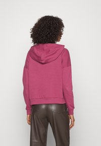 Felpa fucsia corta con cappuccio regolabile, orlo a coste e maniche lunghe. Il modello la indossa con pantaloni in finta pelle marrone.