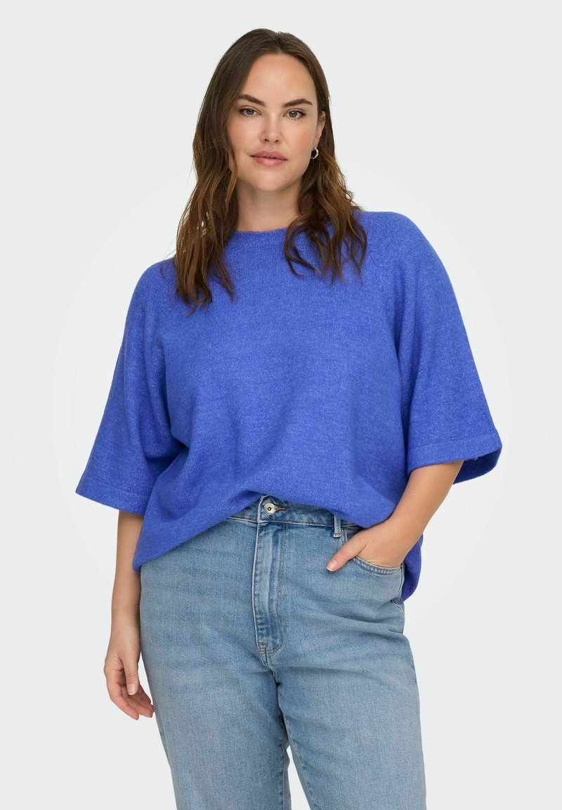 Donna con capelli castani lunghi che indossa un maglione blu a maniche corte e larghe infilato in un paio di jeans a vita alta color azzurro chiaro, con una mano in tasca.