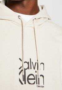 Beige hoodie met een trekkoord, voorzien van een groot zwart "Calvin Klein" logo. Zachte textuur en ribbels aan de mouwen, geschikt voor casual wear.