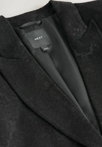 Černý blazer s květinovým vzorem a saténovou podšívkou. Obsahuje klopové límce a štítek s nápisem "NEXT" uvnitř. Elegantní design a látka.