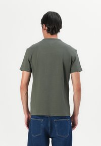 Tricou de culoare verde măsliniu, cu mâneci scurte și guler rotund, având o cusătură în formă de V pe spate, fabricat dintr-un material moale de bumbac. Purtați cu blugi albaștri.