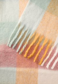 Manta de lã em cores pastel, com um padrão em grelha. Franjinhas texturizadas em amarelo, branco e azul na borda realçam o seu design.