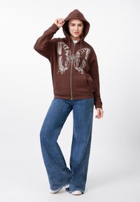 Brūns ar rāvējslēdzēju aiztaisāms džemperis ar lielu sudraba tauriņa grafiku, valkāts kopā ar platiem zilajiem džinsiem. Džemperim ir kapuce ar aukliņu un sānu kabatas.