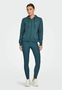 Turkizna zip-up jopica s kapuco in vrvico, v kombinaciji z usklajenimi teksturiranimi legginsami. Bele športne superge zaokrožajo outfit.