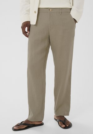 Férfi, bézs színű egyenes szárú nadrágban, fehér ingben, krémszínű kabátban és fekete papucsban, zsebében tartott kézzel áll fehér háttér előtt.