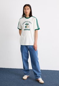White t-shirt with green accents, featuring "ORIGINALS DEPT. 1972" and adidas logo. Paired with relaxed blue jeans and beige sneakers.