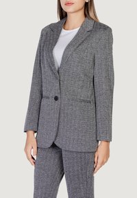 Blazer grigio a motivi a spina di pesce con chiusura a un bottone e rever a scollatura, con tasche laterali e una vestibilità sartoriale. Il materiale sembra testurizzato.