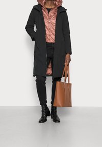 Abrigo largo negro con capucha sobre una chaqueta acolchada color rosa, jeans negros y botas de tacón negras. Bolso tote marrón con dos asas.