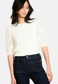 Maglione bianco a maglia con maniche a sbuffo in pizzo. Indossato con jeans in denim scuro a vita alta con chiusura a bottone e tasche posteriori a toppa.
