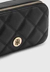 Czarny pikowany portfel wykonany ze skóry teksturowanej z ozdobami w kolorze złotym, wyposażony w okrągły akcent z logo oraz zamek błyskawiczny wzdłuż górnej krawędzi.