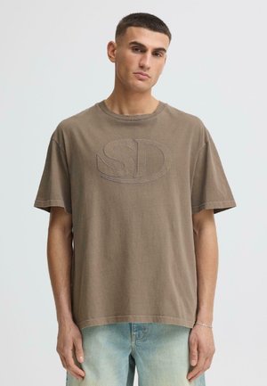 Jonge man met kort donker haar, gekleed in een loszittend bruin T-shirt met "SD" op de voorkant geborduurd en lichtblauwe verweerde spijkerbroek, tegen een witte achtergrond.