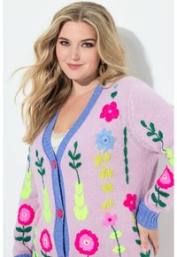 Dzianinowy kardigan w pastelowym różu, ozdobiony żywą, kwiatową haftą w różowym, niebieskim i zielonym kolorze. Z niebieskim kołnierzem i czerwonymi guzikami.