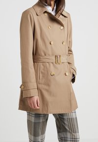 Kvinna i beige dubbelknäppt trenchcoat med bälte och rutiga byxor, händerna i fickorna, mot en enkel ljus bakgrund.