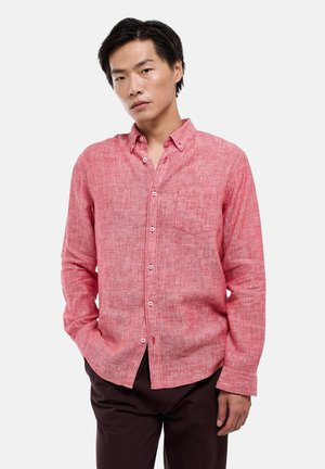 Jeune homme aux cheveux noirs portant une chemise rouge à manches longues boutonnée et un pantalon foncé, debout avec une main dans la poche.