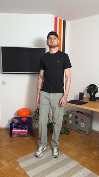 Juoda trumpomis rankovėmis marškinėliai dėvimi su šviesiai žaliais kelnais. Ant rankų matyti tatuiruotės, dėvimi sportbačiai ir kepurė. Vidaus aplinka su spalvingu dekoru.