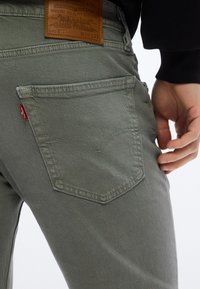 Κοντινό πλάνο στην τσέπη γκρι τζιν Levi’s με καφέ δερμάτινη ετικέτα Levi Strauss και χέρι που ακουμπά κοντά στο ισχίο.