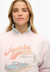 Lichtroze sweatshirt met "Superdry" logo, met een bootafbeelding. Gelaagd met een blauw-witte gestreepte overhemd met kraag eronder.