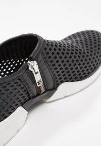 Baskets noires à enfiler avec semelle blanche, dessus perforé et fermeture éclair latérale argentée pour un enfilage facile et une bonne ventilation.