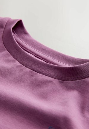 Lilla puuvillane T-särk, millel on ribatud ümardatud kaelus. Kanga tekstuur on sile ja kerge läikega; minimalistlik disain ilma graafikata.