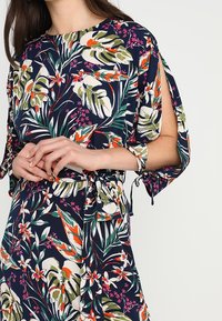 Vestido floral con base azul marino, que presenta patrones tropicales en verde, blanco y naranja. Tiene mangas cortas y un detalle de cintura atada.